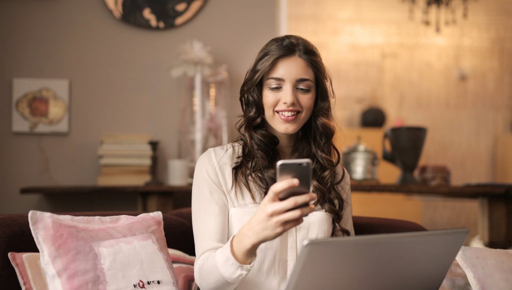 woman sitting on sofa while looking at phone with laptop on lap